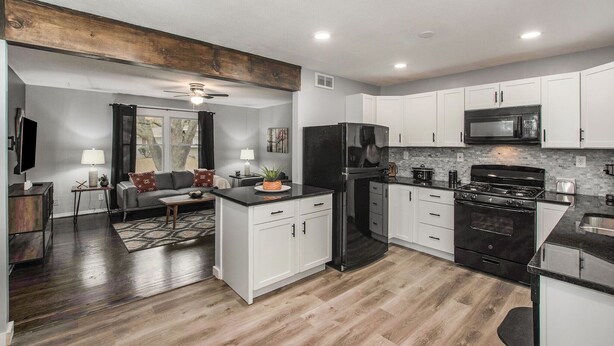 A modern kitchen with white cabinets, a black refrigerator, and a stove. There is a dining area with a table and chairs. - Remodeled 3BR w/ queen beds on 1/2 acre backing to open space. Bonus basement living room + extra bath. Chef’s kitchen, W/D, patio + outdoor dining. Bright, private setting w/ shade trees near Raymore/Belton. Fast WiFi. (Grandview)
