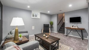 A living room with grey walls, a wooden coffee table, a grey sofa, a black chair, a lamp, a small plant, a TV on a stand, and a staircase leading to another room. - Remodeled 3BR w/ queen beds on 1/2 acre backing to open space. Bonus basement living room + extra bath. Chef’s kitchen, W/D, patio + outdoor dining. Bright, private setting w/ shade trees near Raymore/Belton. Fast WiFi. (Grandview)