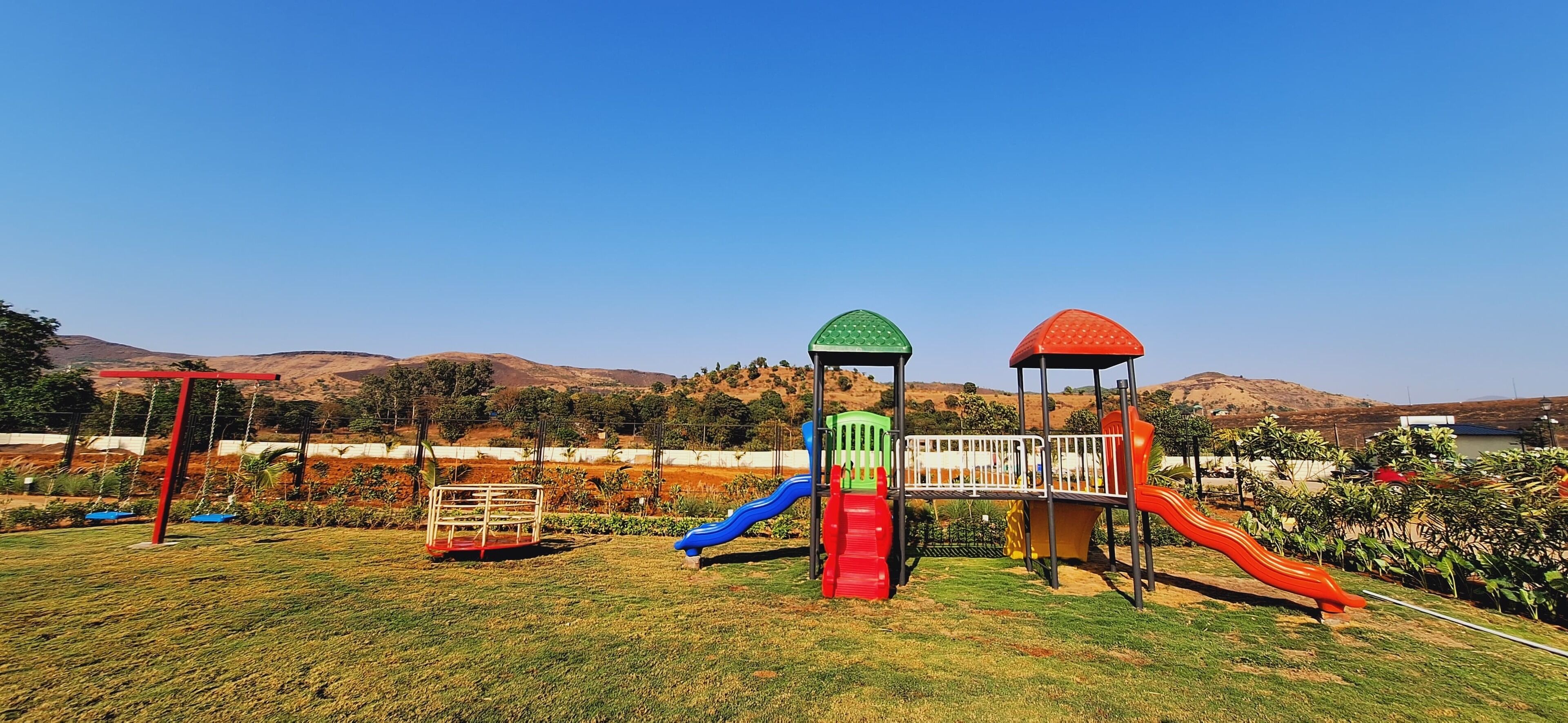 Un parque infantil con toboganes y columpios, rodeado por una valla y colinas al fondo.