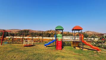 Un parque infantil con toboganes y columpios, rodeado por una valla y colinas al fondo.