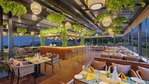 Een restaurant met hoge plafonds, hangplanten en een bar. Tafels zijn gedekt met gele servetten en bestek.