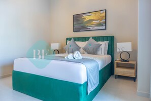 A neatly made bed with a green headboard, white bedding, and two pillows. There is a small bedside table with a lamp and a framed picture on the wall above the bed.