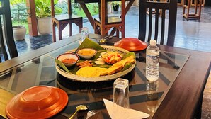 Una mesa de comedor dispuesta con diversos platos, incluyendo un tazón de sopa, una fuente de arroz y una cesta de frutas. Hay sillas alrededor de la mesa, y una botella de agua encima.
