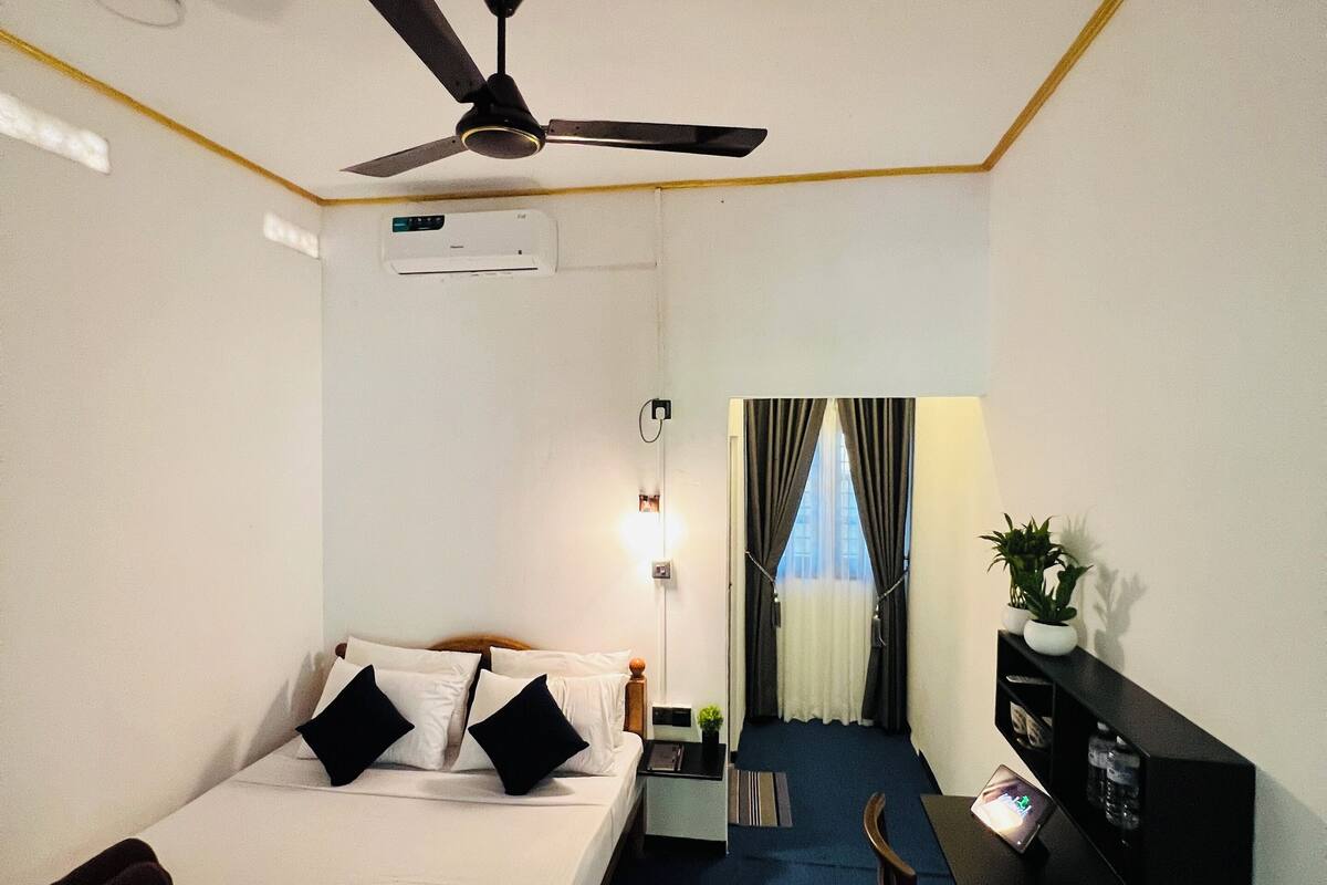 Une chambre d’hôtel équipée d’un lit, d’un bureau, d’une chaise, d’un ventilateur de plafond, d’un appareil de climatisation et offrant une vue sur l’extérieur grâce à une fenêtre avec des rideaux.