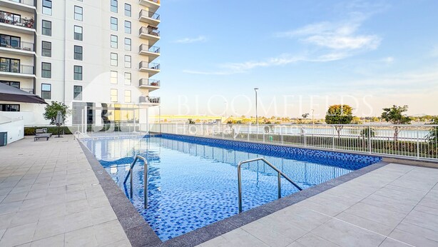 Une piscine dont le bord est revêtu de carreaux, entourée d’une clôture blanche et située à côté d’un immeuble à plusieurs étages doté de balcons.