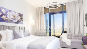 A modern hotel room with a large bed, a sofa, a desk, and a view of the city through large windows. - Bloomfields STU In Al Maryah Island Escape (Abu Dabi)
