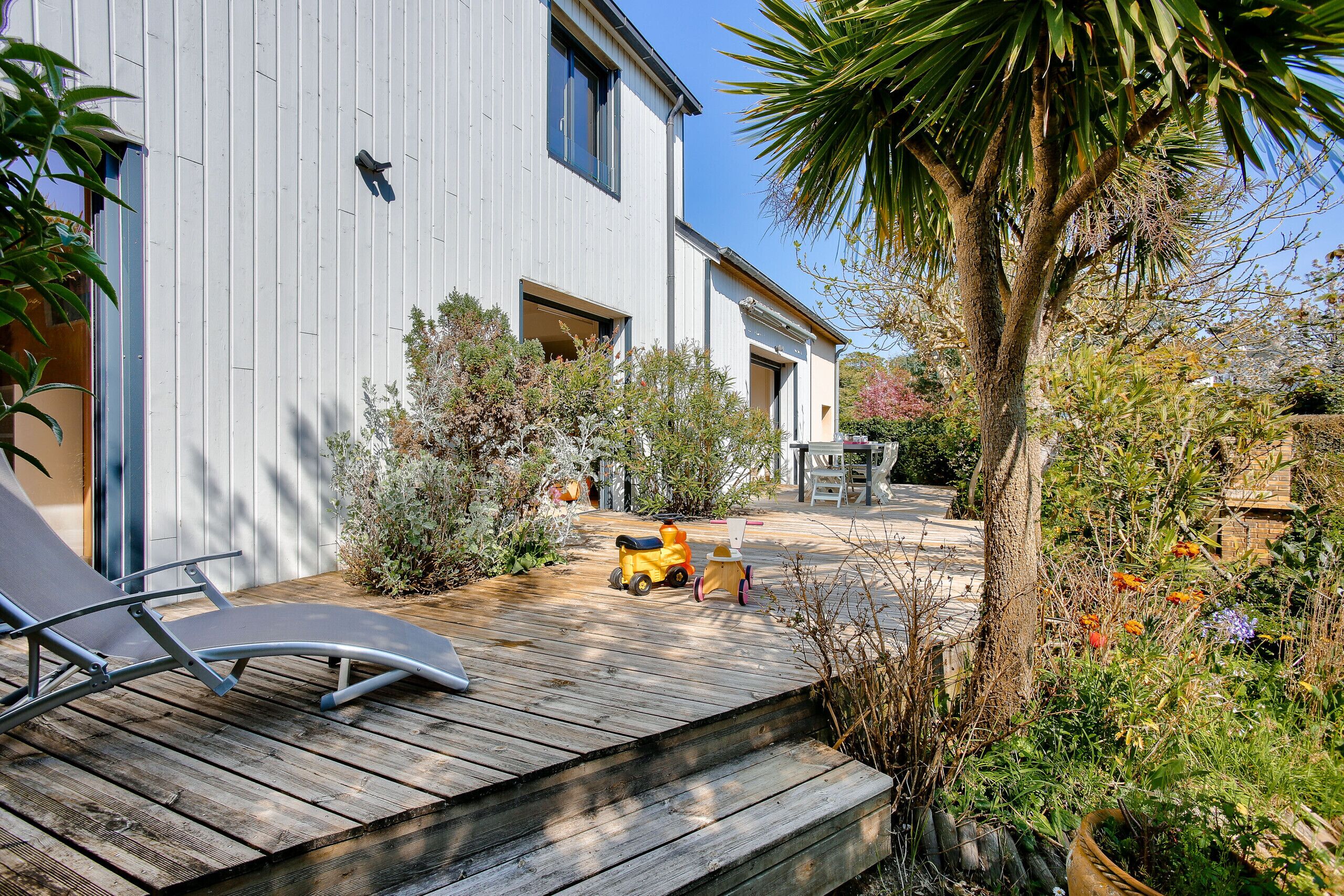 Une maison moderne dotée d’une terrasse en bois, d’un petit jardin et d’un palmier.