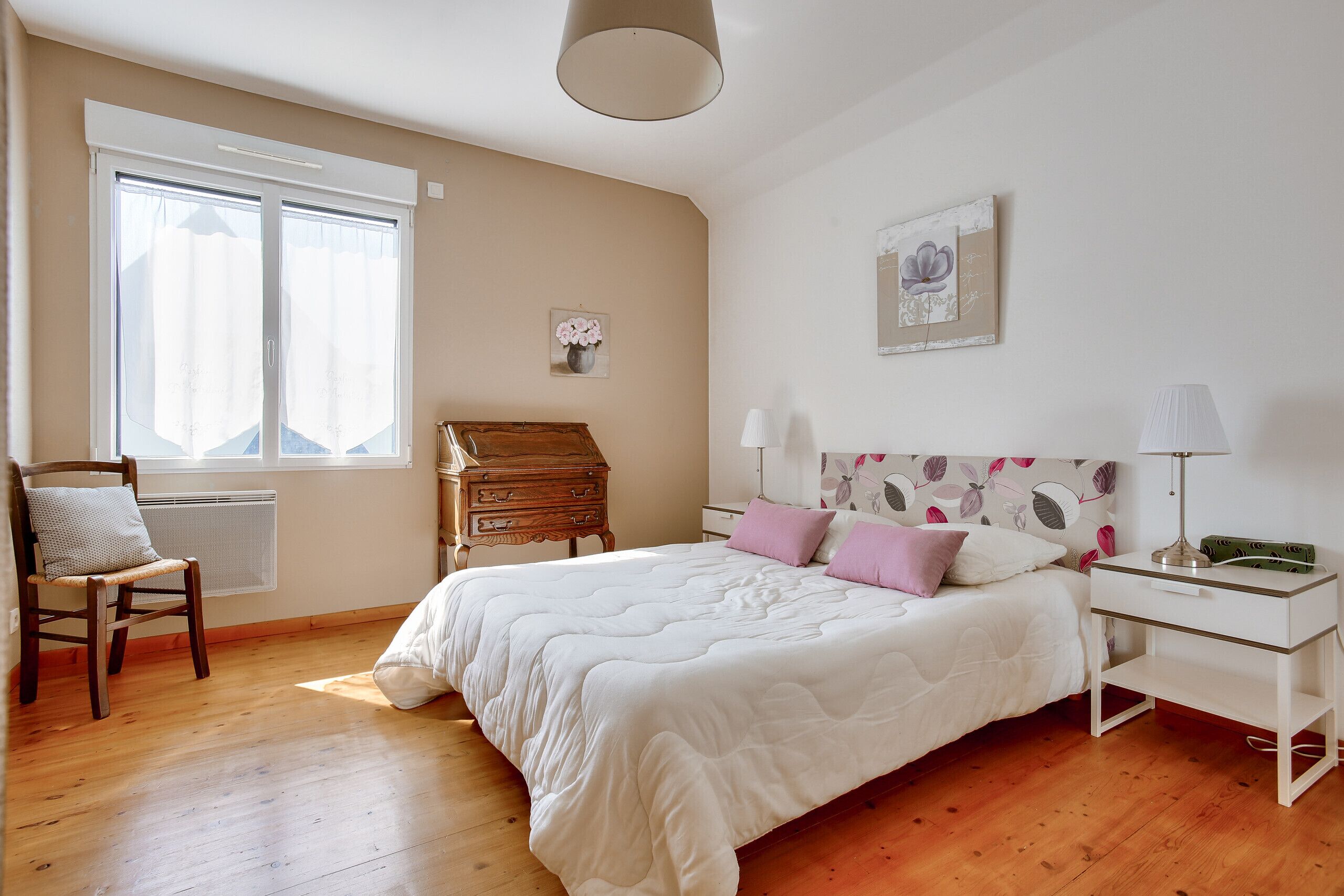 Ein Schlafzimmer mit Holzboden, einem weißen Bett mit pinken und weißen Kissen, einem Holzkapständer, einem weißen Nachttisch mit Lampe und einem Fenster mit Vorhängen.