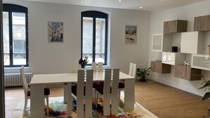 A modern dining area with a white table and chairs, large windows, and a colorful rug. - Modern city-center gîte in Moulins for 4 with Wifi (Moulins)