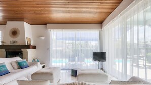 Une chambre d’hôtel moderne avec un plafond en bois, de grandes fenêtres avec des rideaux légers, un téléviseur à écran plat et une vue sur une piscine.