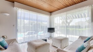 Une pièce lumineuse, avec un plafond en bois, de grandes fenêtres, un téléviseur à écran plat et une vue sur la piscine.