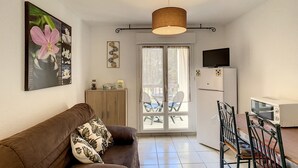 A cozy living room with a brown sofa, a dining table with chairs, a TV mounted on the wall, and a refrigerator.