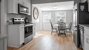 A modern kitchen with grey cabinets, a built-in oven, a microwave, and a refrigerator. There is a dining table with chairs and a clock on the wall. - 3 Blocks to Beach/Downtown & 2 Parking Spots, 3 BLOCKS FROM THE BEACH & BOULEVARD -Lindy's Landing (Carolina Beach)