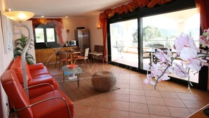 A living room with a sofa, chairs, a table, a TV, a kitchen area, and a balcony with chairs and a table.