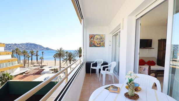 Un balcone con vista sulla spiaggia e sulle montagne, un tavolo da pranzo con una ciotola e un libro, e una televisione nella stanza.