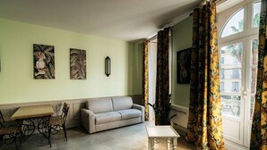A living room with a sofa, a table, and a dining area with chairs. There are two paintings on the wall and a window with curtains. - Charming air-conditioned apartment, central Pézenas, pets welcome (Pézenas)