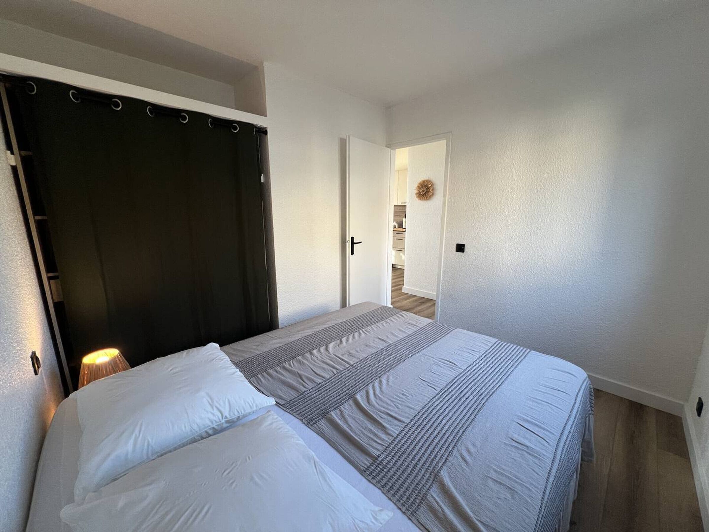 Un dormitorio con una cama, cabecera oscura, lámpara de noche y una puerta que da a un pasillo.