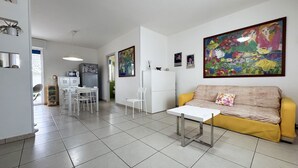 Una sala de estar moderna con un sofá amarillo, una mesa de centro blanca y un comedor con una mesa y sillas blancas. En la cocina hay dos refrigeradores, un microondas y un fregadero.