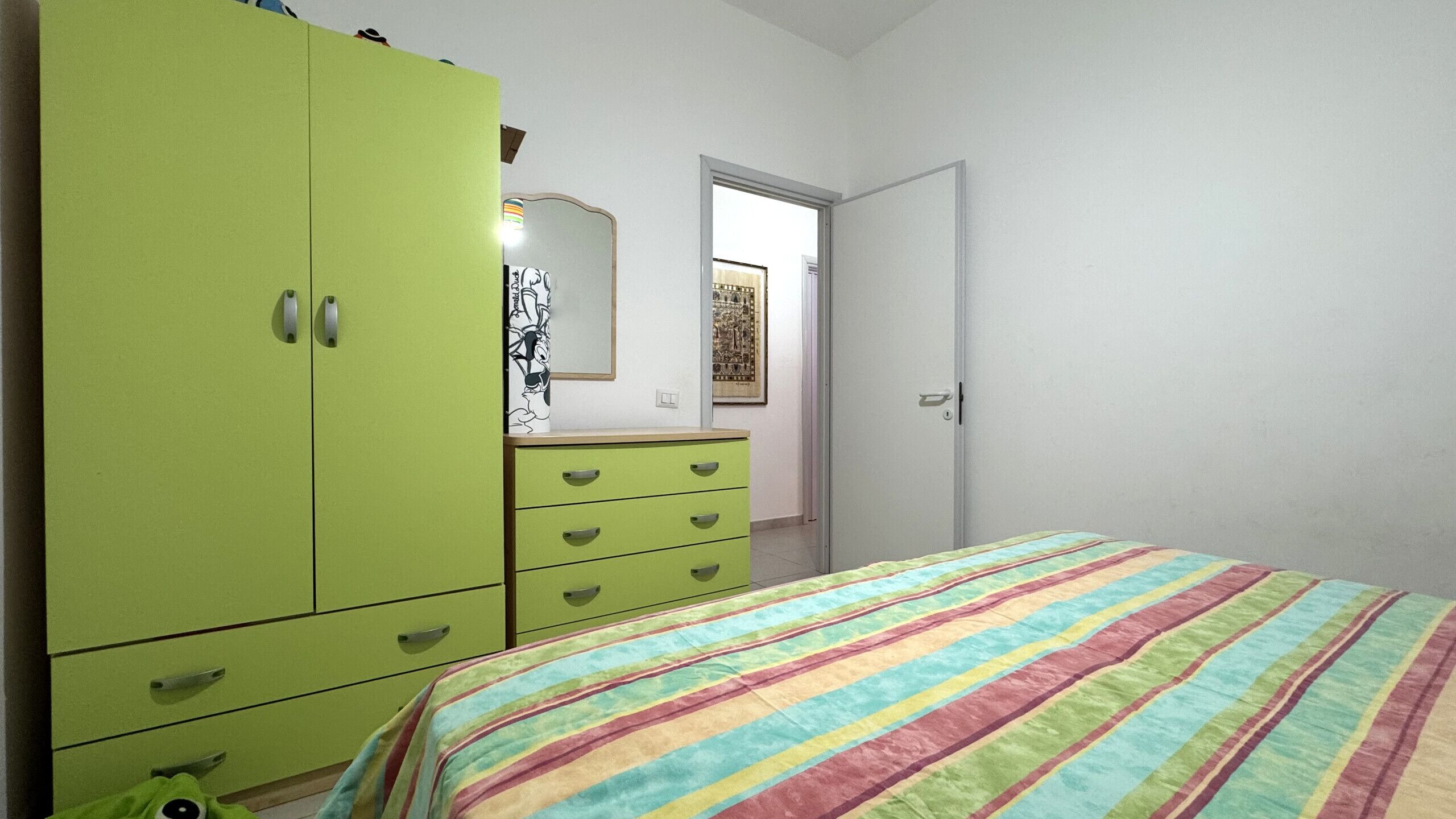A bedroom with a green wardrobe, a bed with a striped blanket, and a door leading to another room.