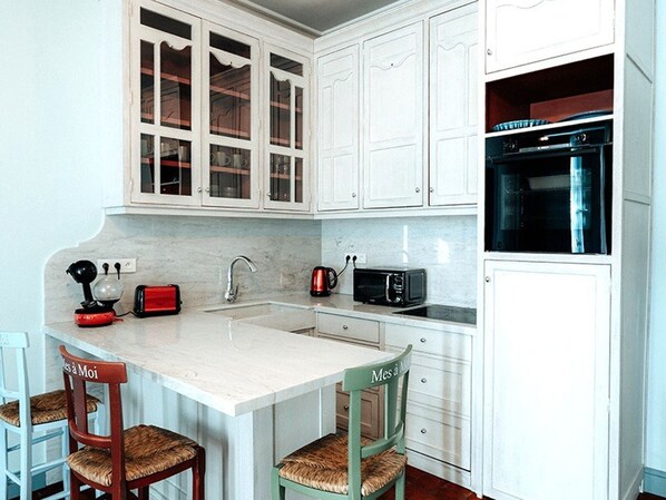 A kitchen with white cabinets, a marble countertop, a sink, a kettle, a toaster, and a microwave. There are two wooden chairs with woven seats.