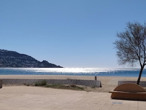 A paved walkway leading to a beach with a clear view of the sea and a hill with buildings in the background.