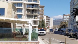 A multi-story residential building with balconies, a cafe with outdoor seating, and parked cars on the street. - Modern 2-bedroom apartment with private garden 100m from beach (Roses)
