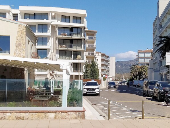 A multi-story residential building with balconies, a cafe with outdoor seating, and parked cars on the street.