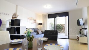 A modern living room with a sofa, a dining table with plates and glasses, a TV mounted on the wall, and a view of the outdoors through a large window. - Modern 2-bedroom apartment with private garden 100m from beach (Roses)