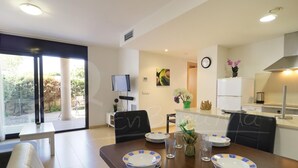 A modern dining area with a wooden table set for four, a TV mounted on the wall, and a kitchen area with a refrigerator and stove. - Modern 2-bedroom apartment with private garden 100m from beach (Roses)