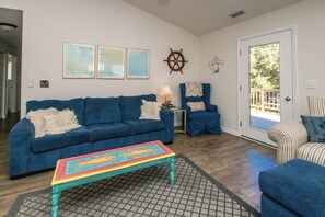 A living room with a blue sofa, a colorful coffee table, and a striped chair. There are framed pictures on the wall and a ship's wheel decoration.