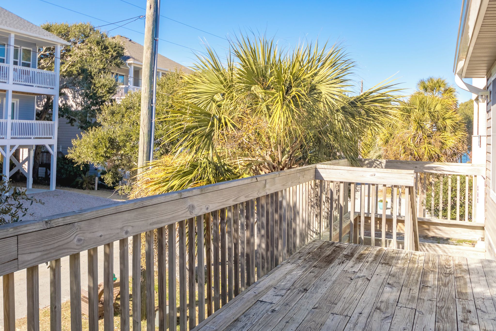 A wooden deck with a railing, a house with a balcony, and a palm tree in the background.