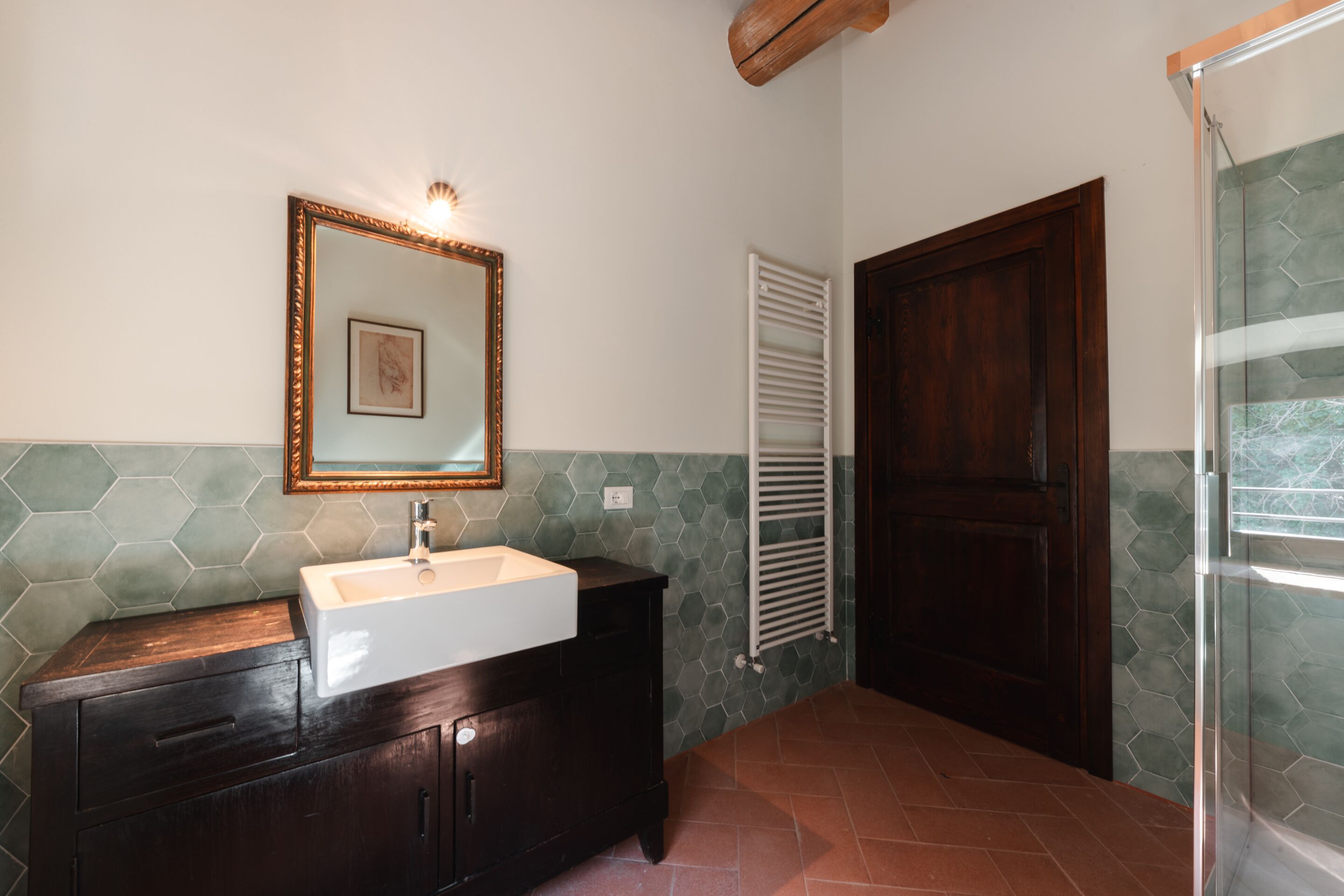 A bathroom with a dark wooden vanity, a white sink, a framed mirror, a hexagonal tiled wall, a white towel rack, and a glass shower enclosure.