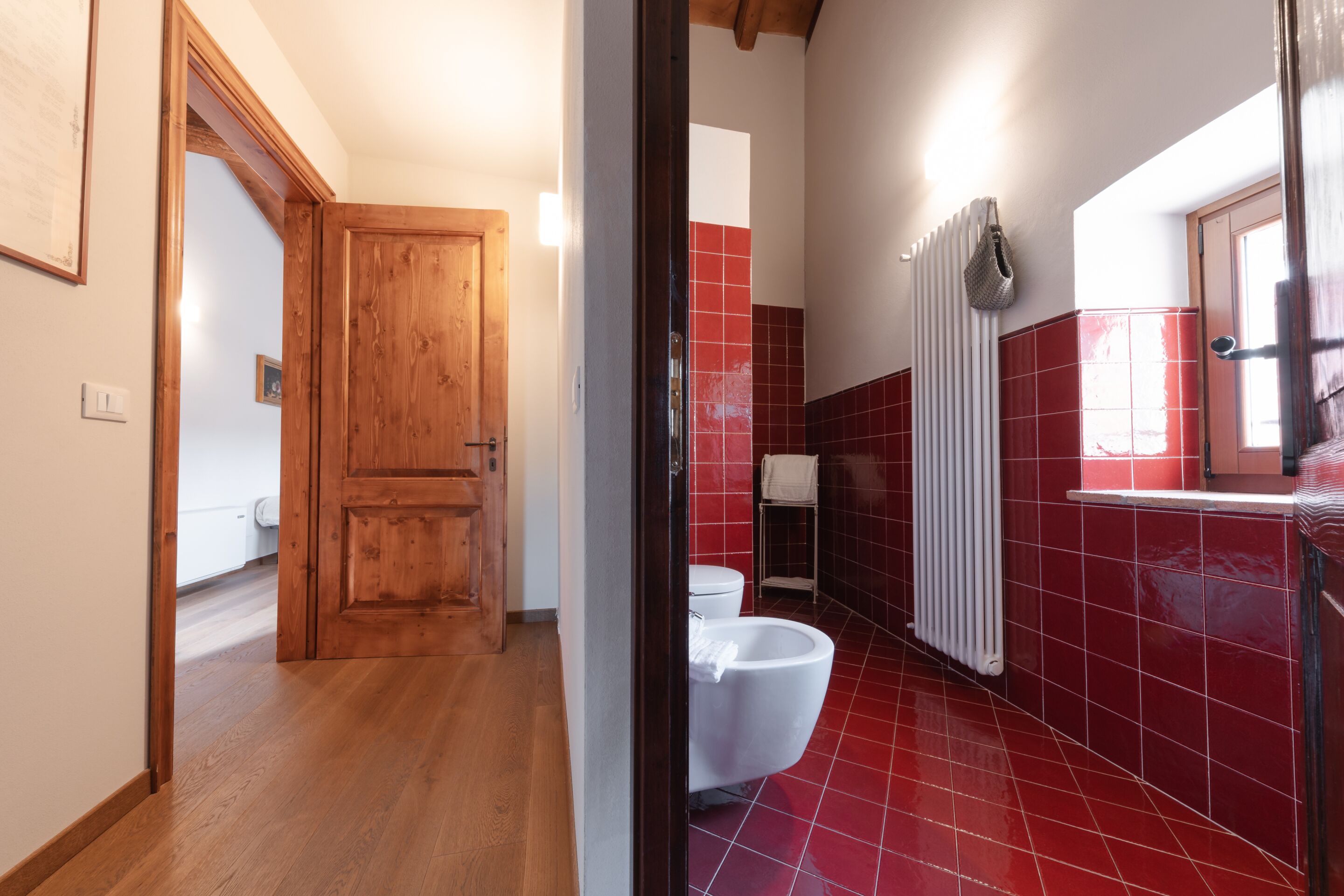 Ein Badezimmer mit Holztür, Toilette in Weiß, Duschbereich mit roten Fliesen und einem weißen Heizkörper.