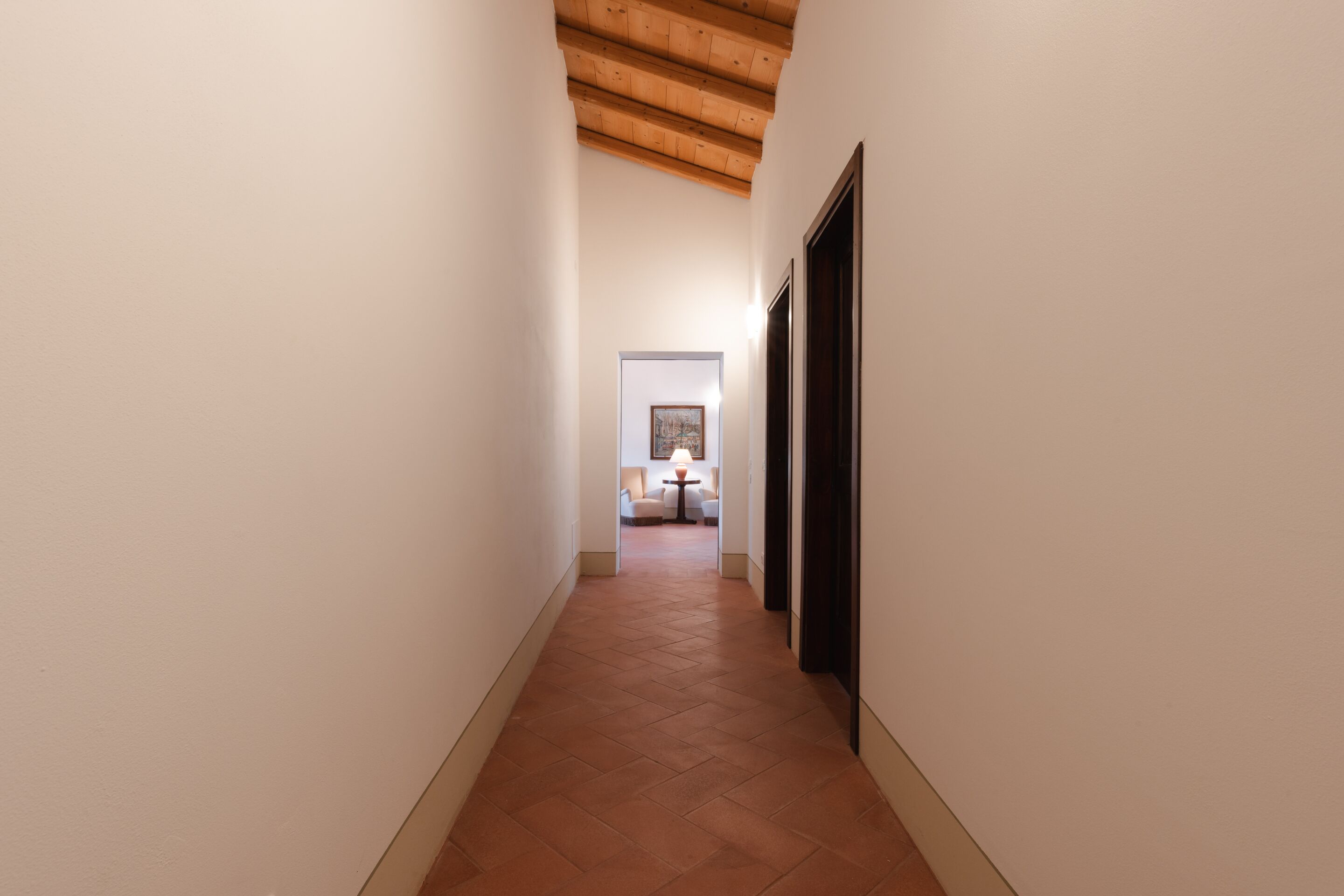 A narrow hallway with a wooden ceiling and a tiled floor leading to a room with a table and chairs.