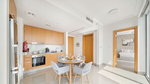 A modern kitchen with wooden cabinets, a dining area with a round table and chairs, and a bedroom visible through an open door.