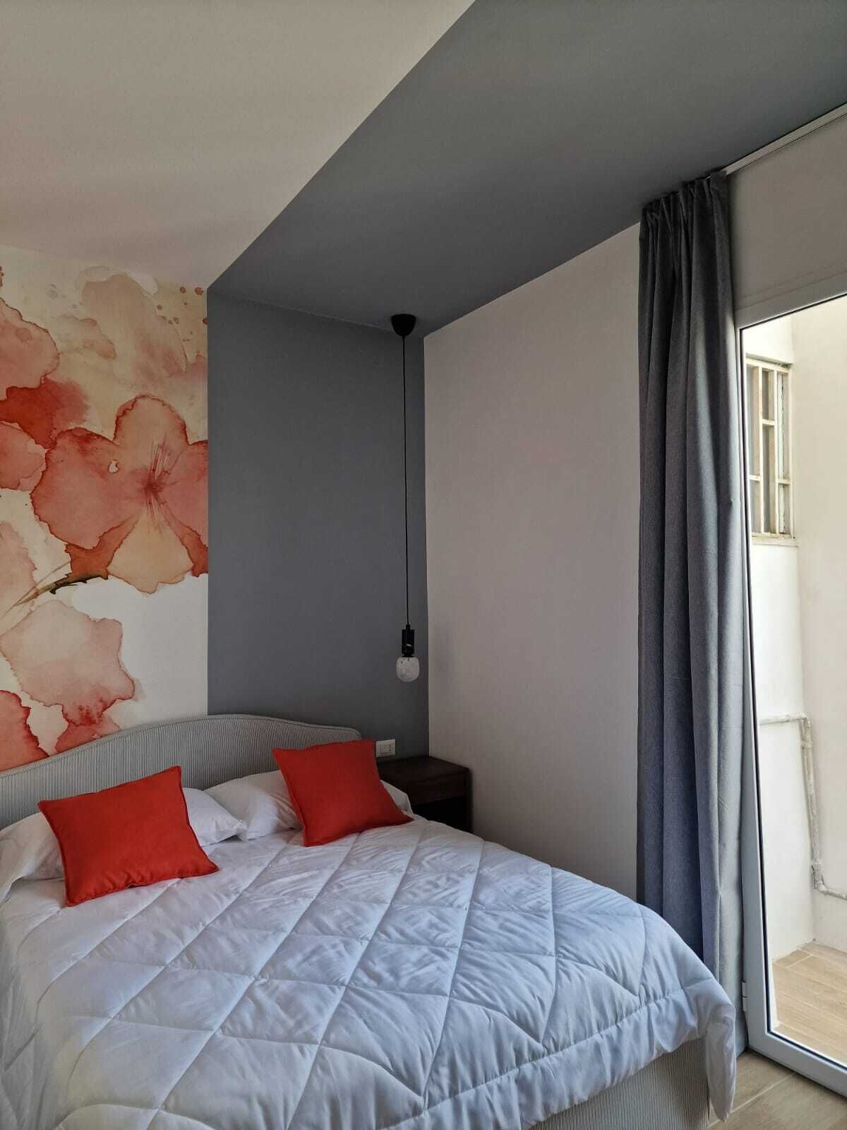 Une chambre à coucher comprenant un lit, une table de chevet, une lampe fixée au mur et une grande fenêtre avec des rideaux.