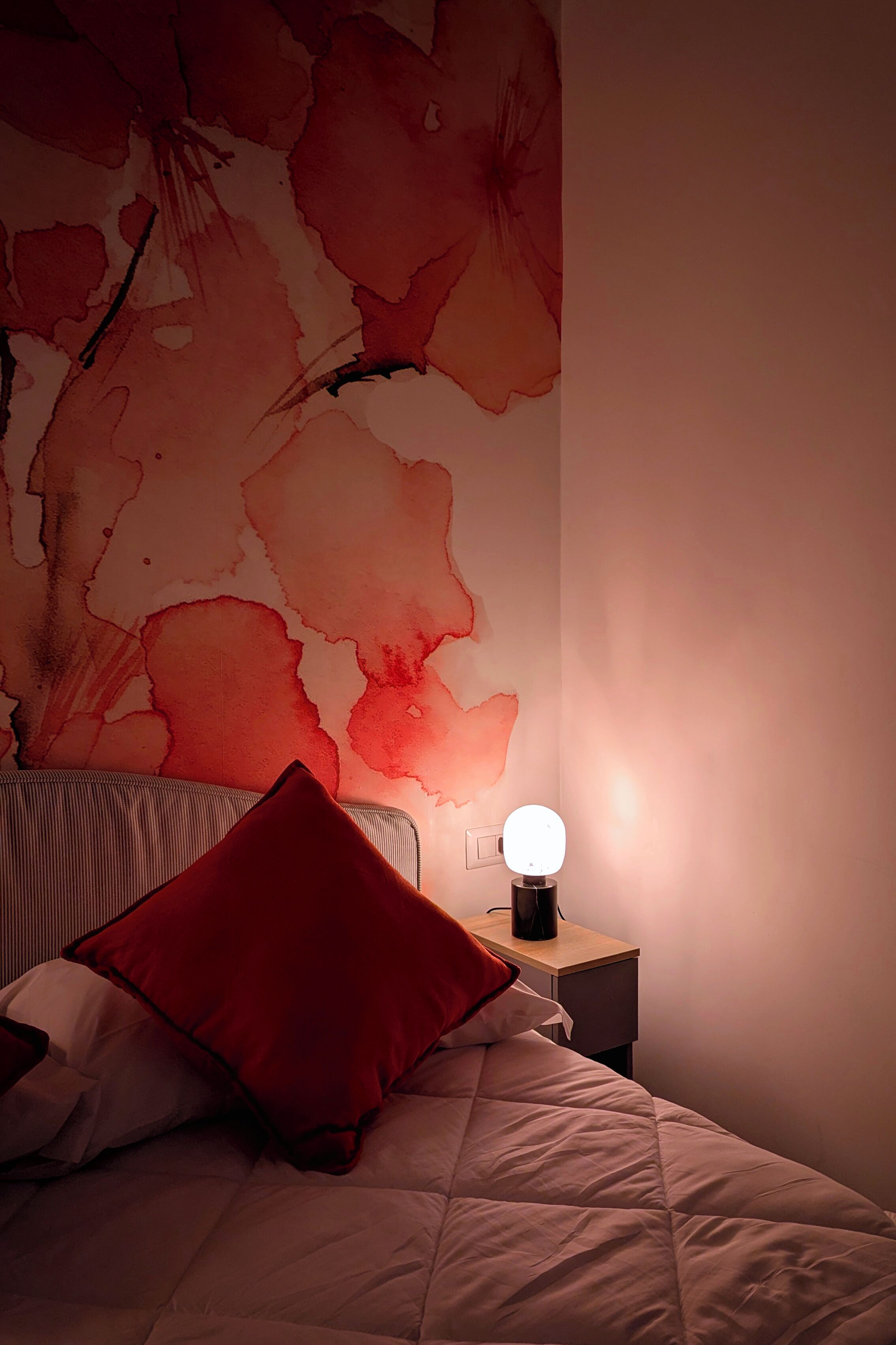 A bedroom with a bed, a nightstand, a wall-mounted lamp, and a floral wallpaper.