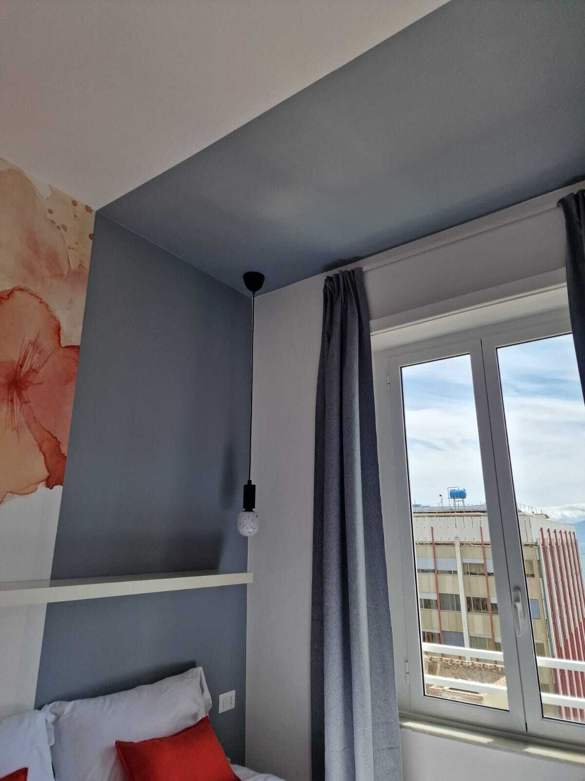 A bedroom with a bed, a window with curtains, a wall with a floral painting, and a ceiling light.