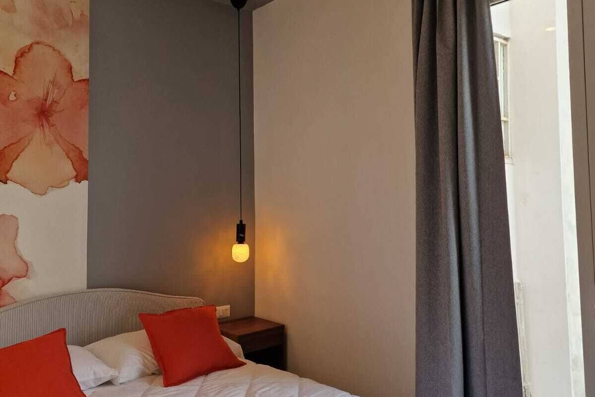 A bedroom with a bed, pillows, a nightstand, a wall with a floral design, a hanging light, and a view through a window with curtains.