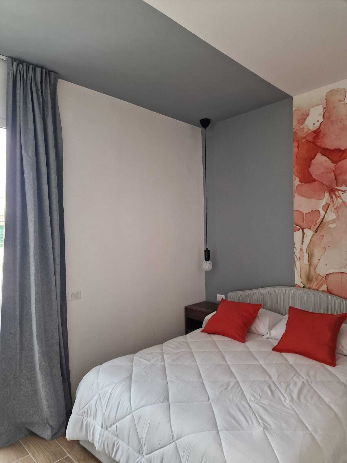 A bedroom with a bed, grey curtains, a nightstand, and a wall with a floral painting.