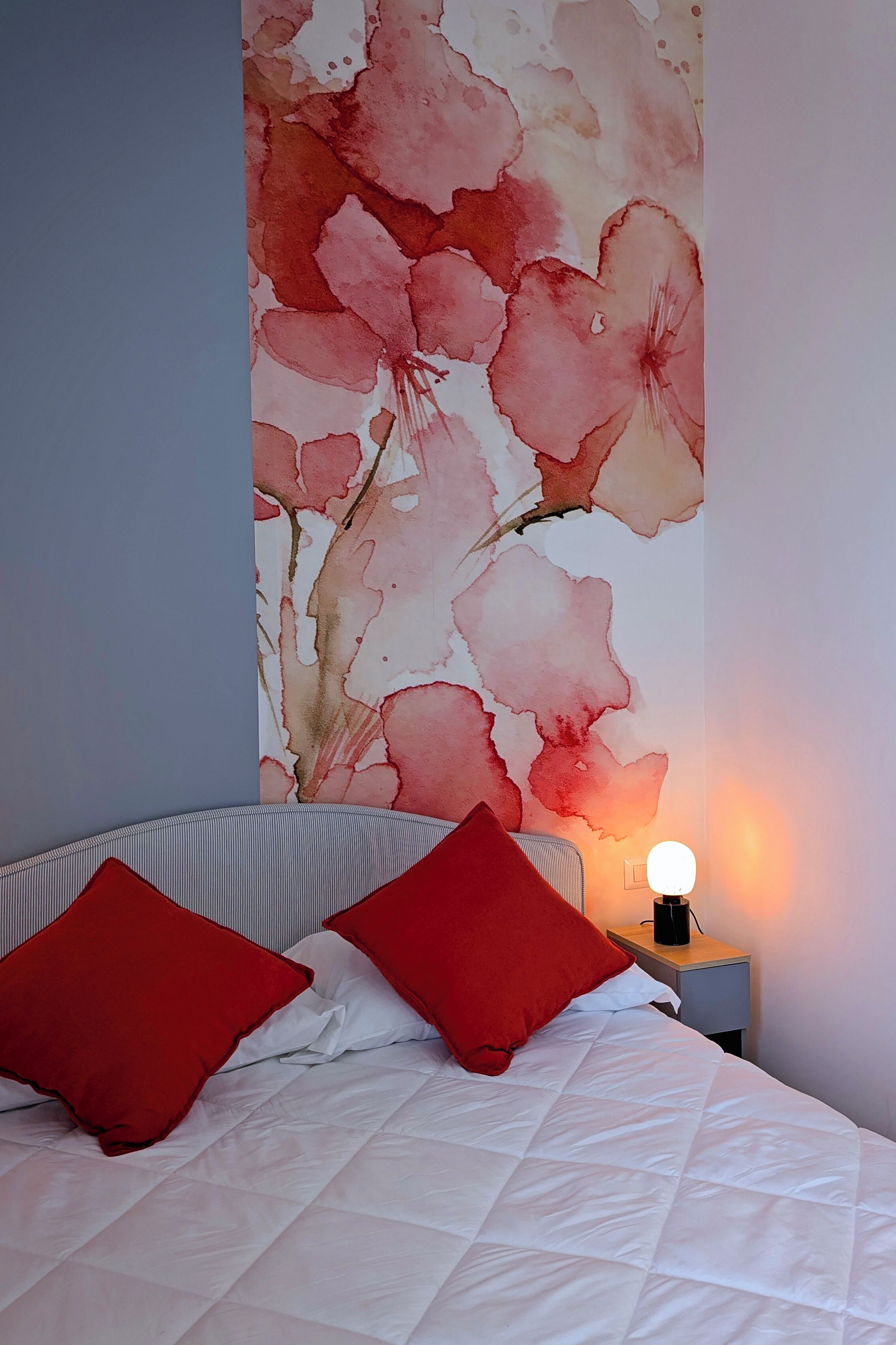 A bedroom with a bed, red pillows, a nightstand with a lamp, and a floral wallpaper.