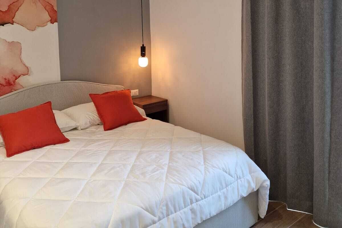 A bedroom with a bed, pillows, a nightstand, a wall with a floral painting, a hanging light, and a window with curtains.