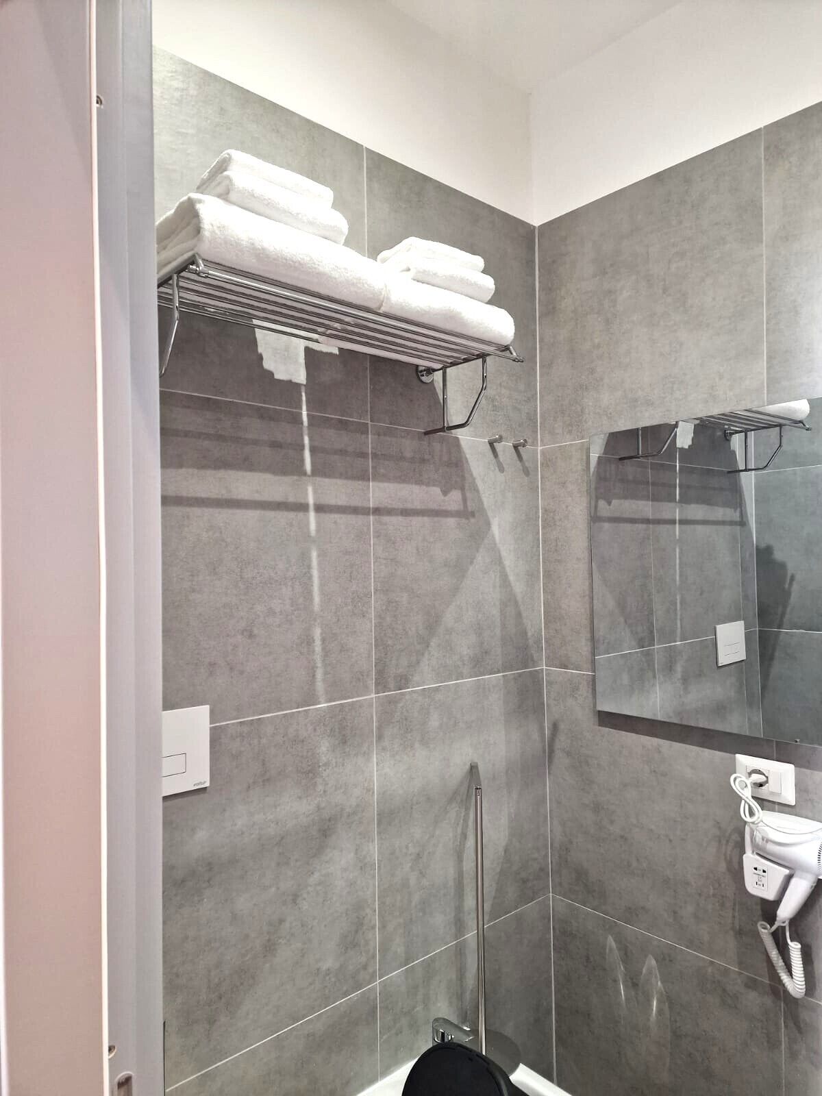 A modern bathroom with grey tiles, a glass shower enclosure, a towel rack with folded towels, a hairdryer, and a mirror.