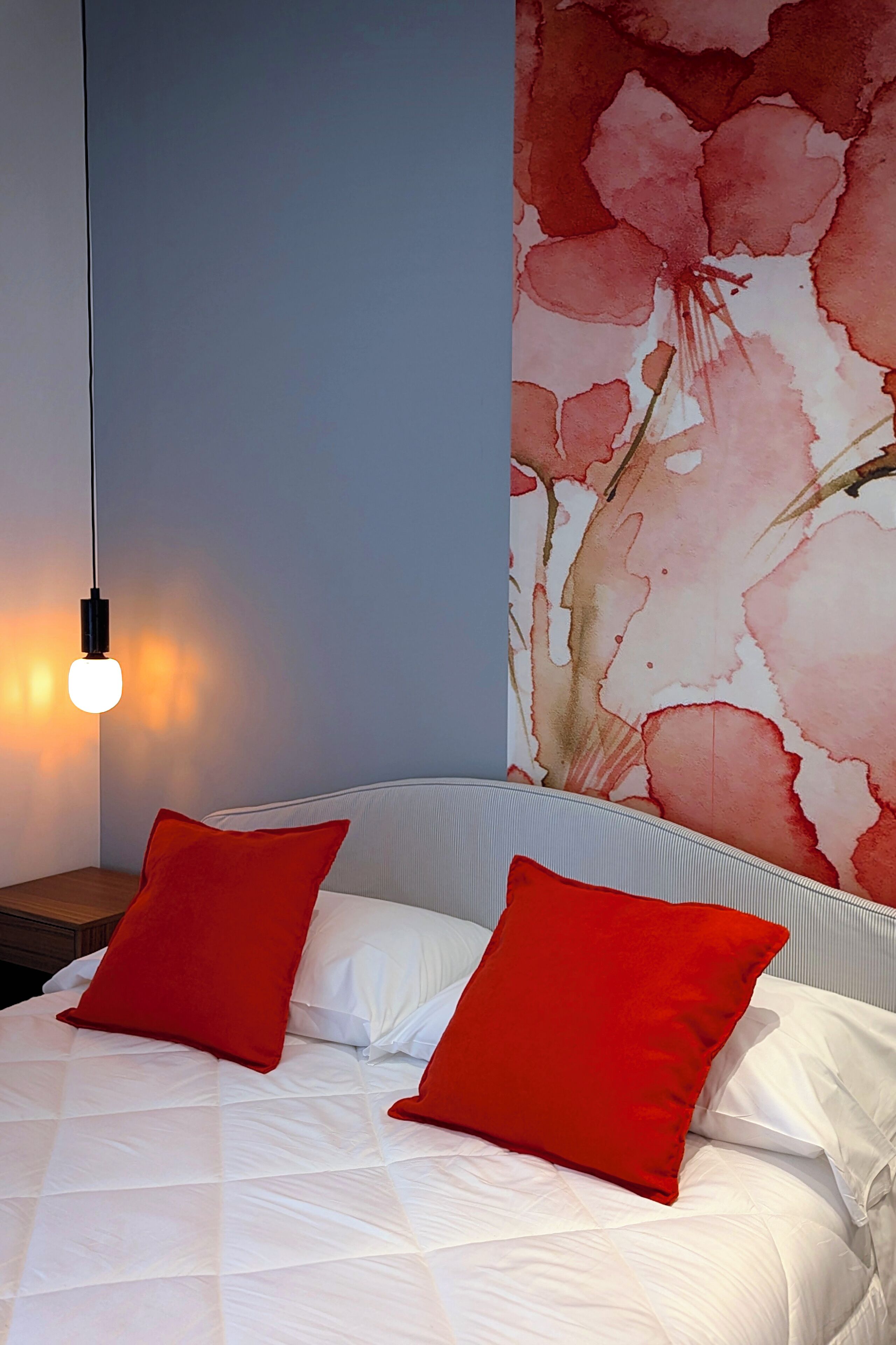 Un lit avec une literie blanche et deux oreillers rouges, un mur orné d’un motif floral et un luminaire suspendu.
