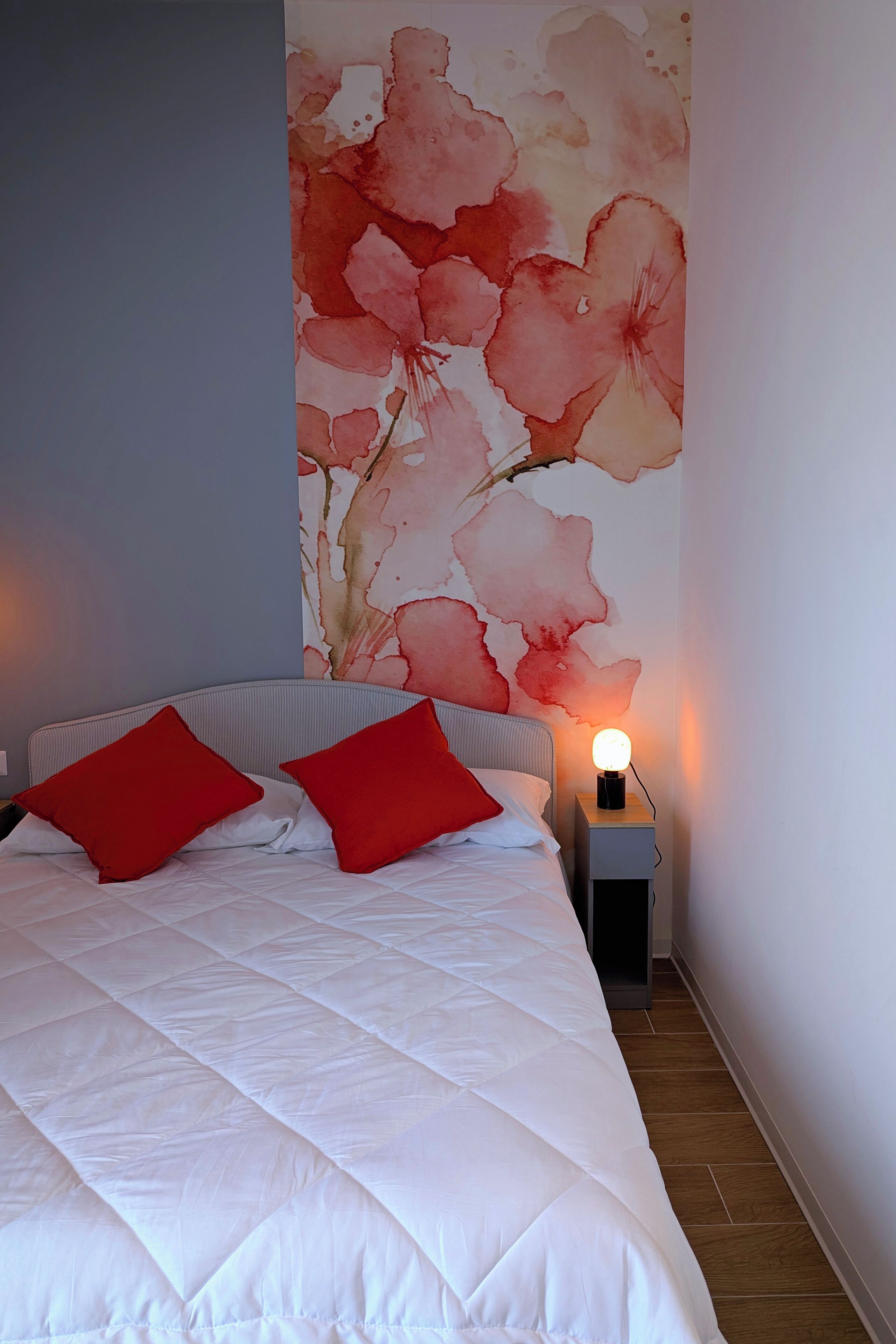 A neatly made bed with a white duvet and two red pillows, a small bedside table with a lamp, and a large floral wall mural.