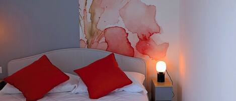Un lit bien fait, avec une couette blanche et deux oreillers rouges, une petite table de chevet avec une lampe, et un grand décor mural représentant des motifs floraux.