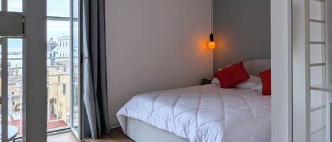 A bedroom with a bed, a window with curtains, a hanging light, and a view of buildings outside.
