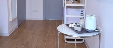 Una habitación con una televisión blanca empotrada en la pared, un estante blanco con escalera, una mesa redonda blanca con una tetera y un control remoto, y suelo de madera.