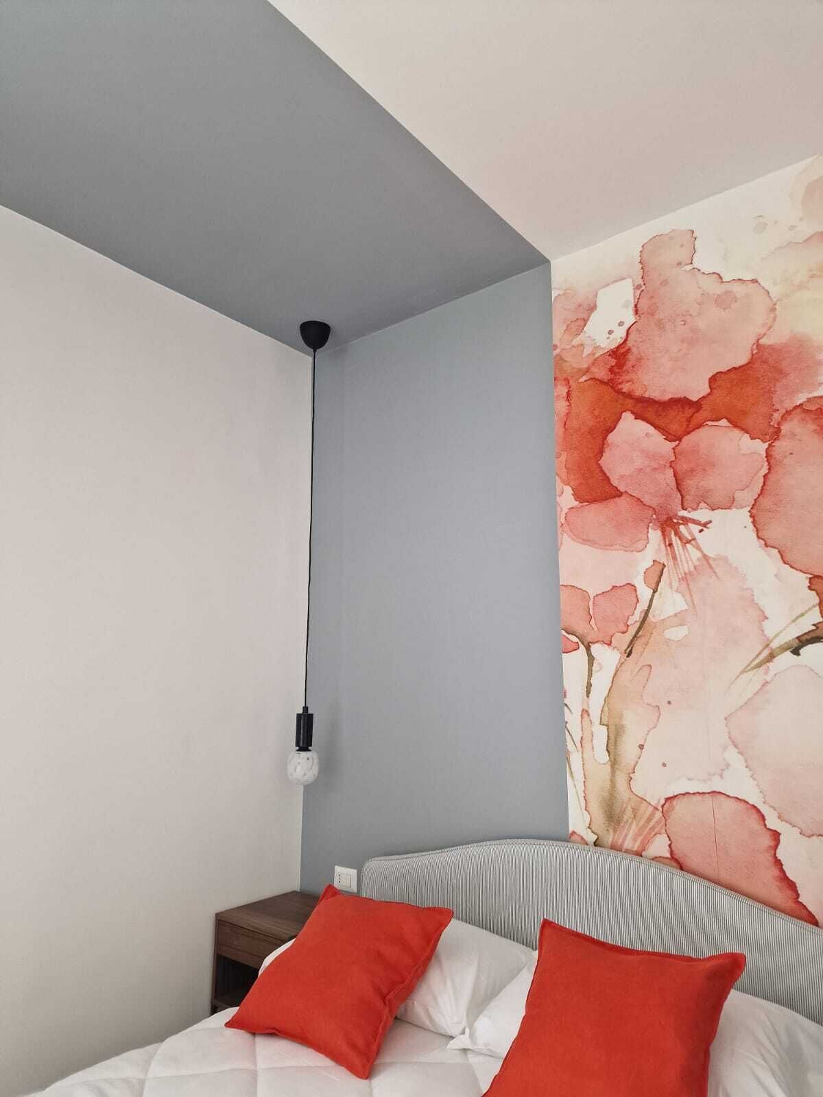 A bedroom with a bed, two red pillows, a nightstand, a wall-mounted light, and a large floral artwork.