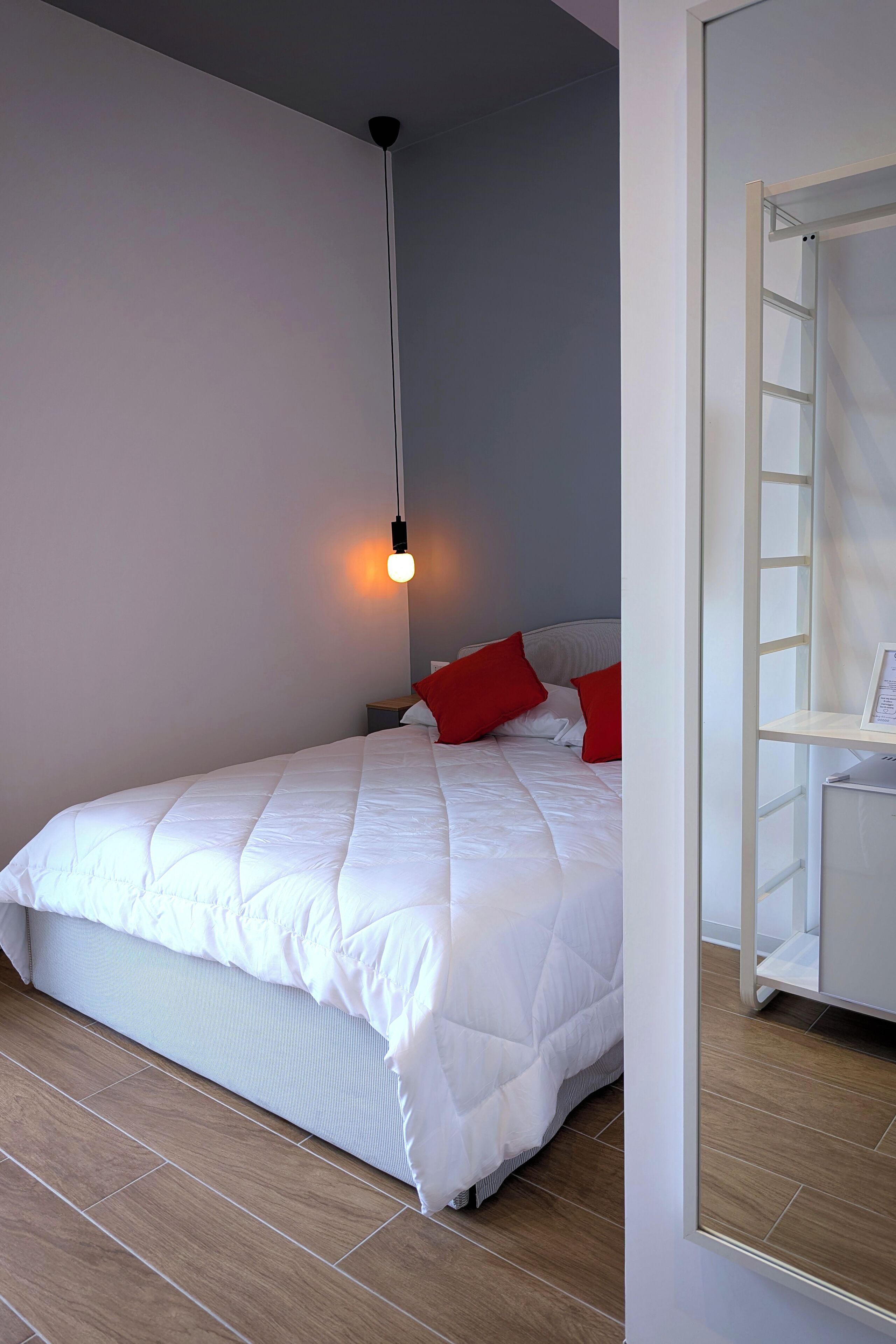 Una cama bien hecha con ropa de cama blanca y almohadas rojas, una lámpara colgante y un armario blanco con una escalera.